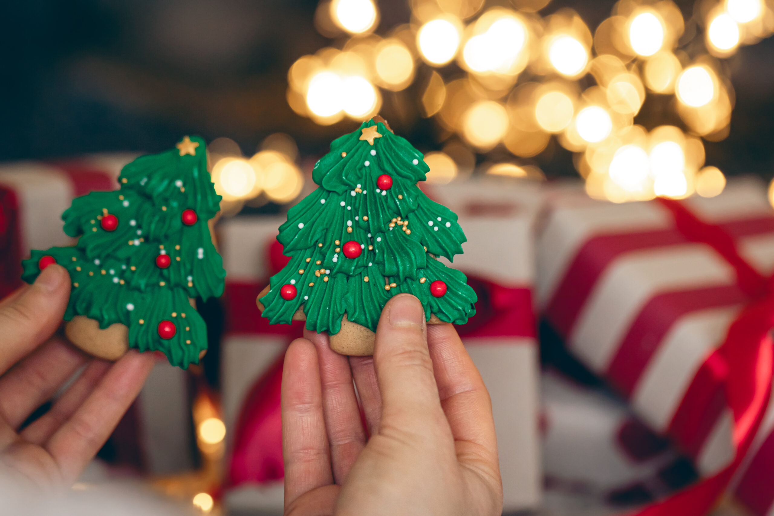 Manos sosteniendo galletas decoradas con forma de árbol de Navidad frente a luces cálidas